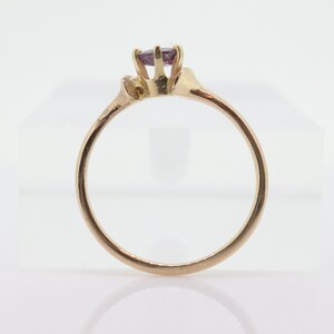May include: A gold ring featuring a small, round, purple gemstone. The ring has a delicate band and a simple setting. The ring is displayed on a white surface.
