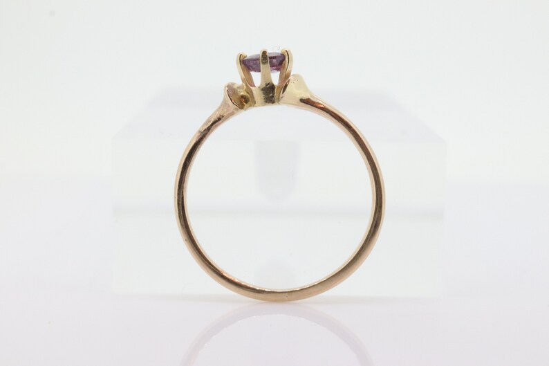 May include: A gold ring featuring a small, round, purple gemstone. The ring has a delicate band and a simple setting. The ring is displayed on a white surface.