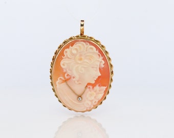 Puede incluir: Un colgante de oro con un camafeo que representa el perfil de una mujer con un pequeño acento de diamante. El camafeo está engastado en un marco de oro con un diseño de cuerda.