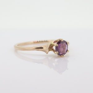 May include: A gold ring featuring a round, purple gemstone. The ring has a plain band and a six-prong setting. The ring is shown on a white surface, reflecting the ring's design. The gemstone is a deep shade of purple.