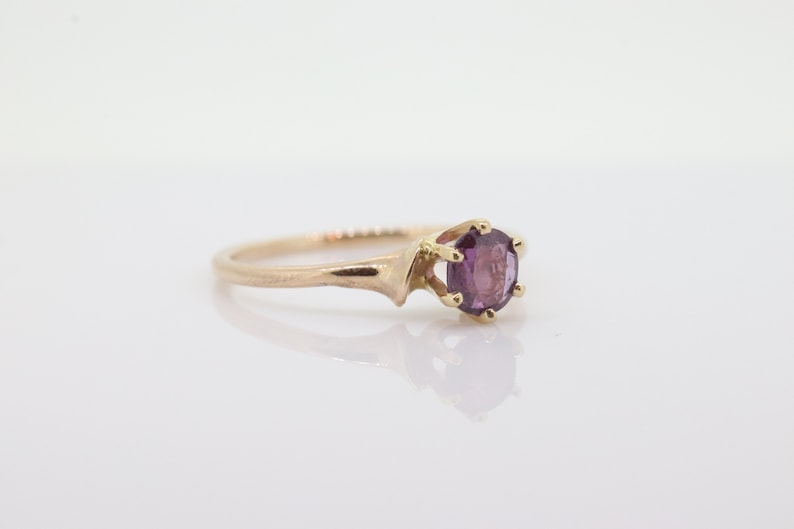 May include: A gold ring featuring a round, purple gemstone. The ring has a plain band and a six-prong setting. The ring is shown on a white surface, reflecting the ring's design. The gemstone is a deep shade of purple.