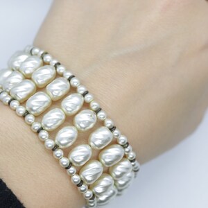 May include: A white pearl bracelet with three rows of large, round pearls. The pearls are strung on a black cord.