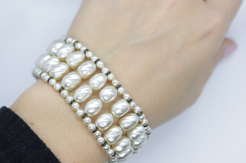 May include: A white pearl bracelet with three rows of large, round pearls. The pearls are strung on a black cord.
