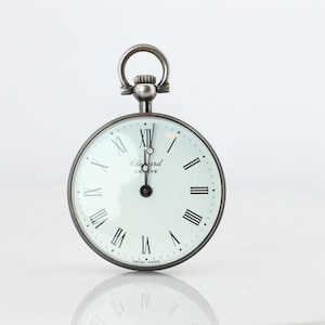 Könnte beinhalten: Eine silberfarbene Taschenuhr mit weißem Zifferblatt und schwarzen römischen Ziffern. Die Uhr hat eine Schlaufe oben und die Worte "Chopard Geneve" sind auf dem Zifferblatt aufgedruckt. Die Uhr spiegelt sich in der darunter liegenden Oberfläche.