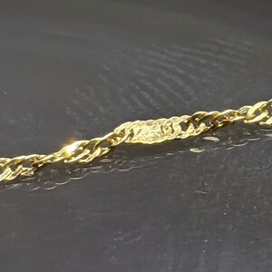 May include: Close-up of a gold-colored chain necklace. The chain has a twisted rope design with a textured rectangular accent piece in the center. The necklace is displayed against a dark background.