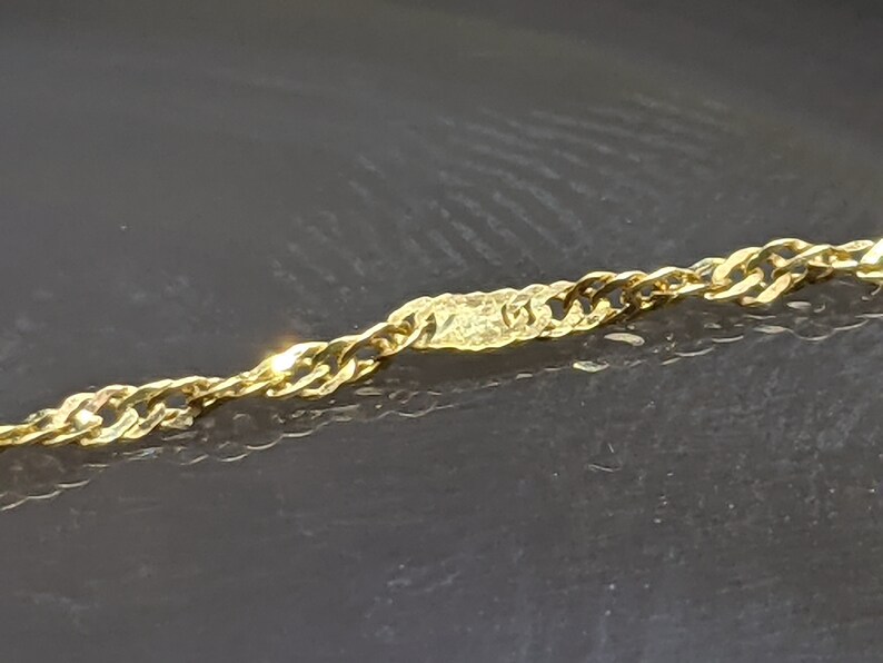 May include: Close-up of a gold-colored chain necklace. The chain has a twisted rope design with a textured rectangular accent piece in the center. The necklace is displayed against a dark background.