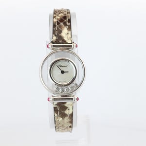 May include: A silver-tone wristwatch featuring a round white dial with the Chopard logo, surrounded by a clear circular frame containing five floating round-cut diamonds. The watch has a snakeskin-patterned strap and small red accents at the lugs.