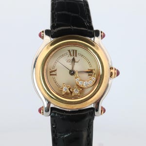 May include: A Chopard wristwatch with a black alligator-textured leather strap, a round silver and gold-tone case, and a cream dial. The dial features Roman numerals, a floating diamond-encrusted crescent moon, and two floating diamond stars.
