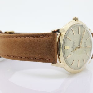 Pu&ograve; includere: Un orologio da polso dorato con quadrante bianco e lancette nere. L'orologio ha un cinturino in pelle marrone.