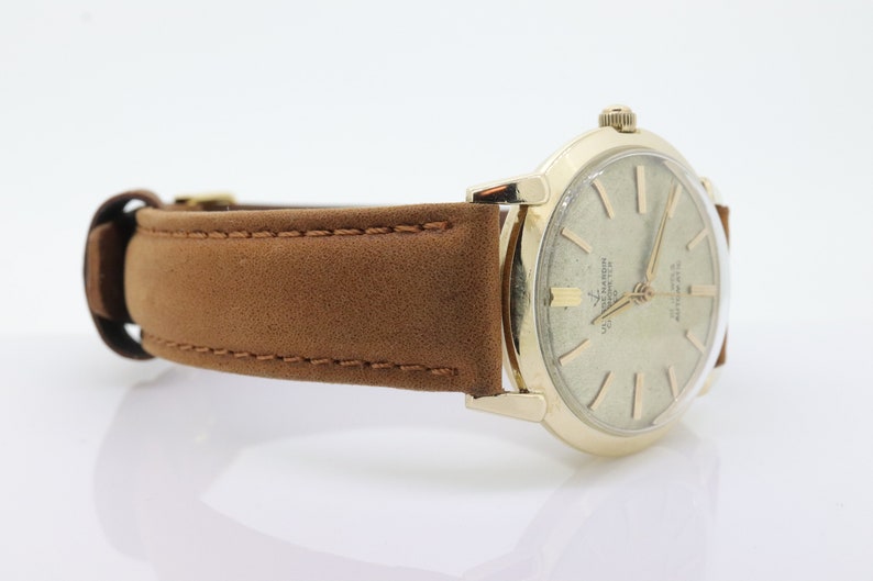 Pu&ograve; includere: Un orologio da polso dorato con quadrante bianco e lancette nere. L'orologio ha un cinturino in pelle marrone.