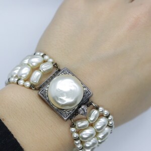May include: A white pearl bracelet with a silver square clasp featuring a large pearl in the center. The bracelet is made of multiple strands of pearls.