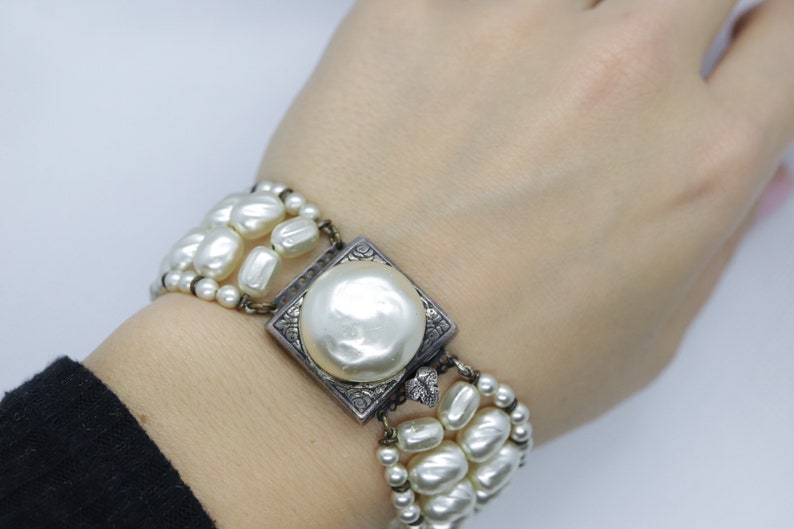 May include: A white pearl bracelet with a silver square clasp featuring a large pearl in the center. The bracelet is made of multiple strands of pearls.