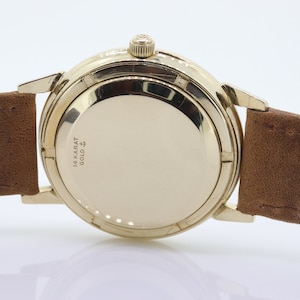 Pu&ograve; includere: Un orologio da polso dorato con un cinturino in pelle marrone. Il retro della cassa dell'orologio &egrave; inciso con "14 KARAT GOLD" e un simbolo.