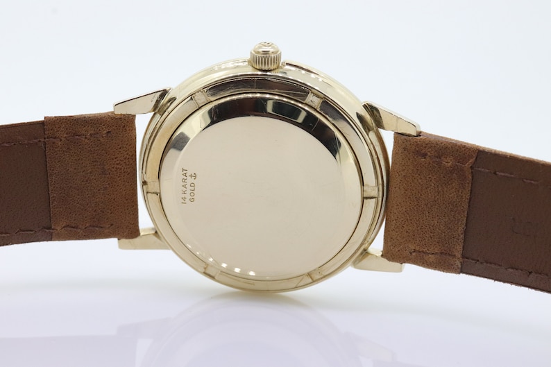 Pu&ograve; includere: Un orologio da polso dorato con un cinturino in pelle marrone. Il retro della cassa dell'orologio &egrave; inciso con "14 KARAT GOLD" e un simbolo.