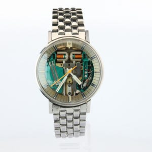May include: A silver-tone Bulova Accutron wristwatch with a round case and a multi-link metal bracelet. The dial features a visible green circuit board movement, white hour markers, and silver hands with a yellow second hand.