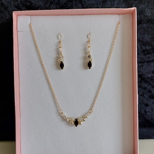 May include: A gold necklace with a delicate chain and a pendant featuring a cluster of small pearls and a dark red gemstone. The necklace is displayed in a pink gift box with matching earrings featuring a single dark red gemstone.