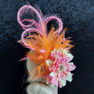 Op de afbeelding: Een opvallend hoofddeksel met een grote roze en crèmekleurige bloem, aangevuld met oranje veren en elegante roze veerslingers. Kleine gouden kralen zorgen voor glamour. Ideaal voor evenementen.