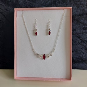May include: A silver necklace and earring set presented in a pink box. The necklace and earrings feature clear and red crystals. The necklace has a central marquise-shaped red crystal. The earrings have a similar design, with a red crystal.