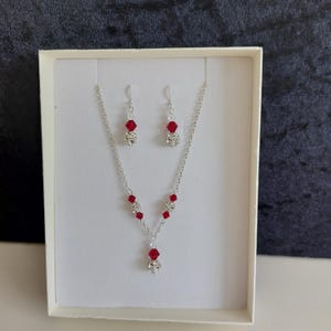 May include: A silver necklace and earrings set with red and clear crystal beads. The necklace has a pendant with a larger red crystal.