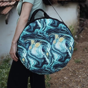 Pode incluir: Uma bolsa redonda de tecido marmoreado em azul-petróleo, preto e dourado com uma alça preta. A bolsa está sendo carregada por uma pessoa.