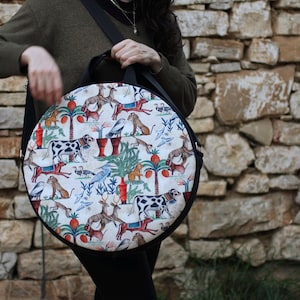 May include: A round, black shoulder bag with a whimsical print featuring cows, deer, birds, and palm trees on a white background. The bag has a black strap and handle. The bag is held in front of a stone wall.