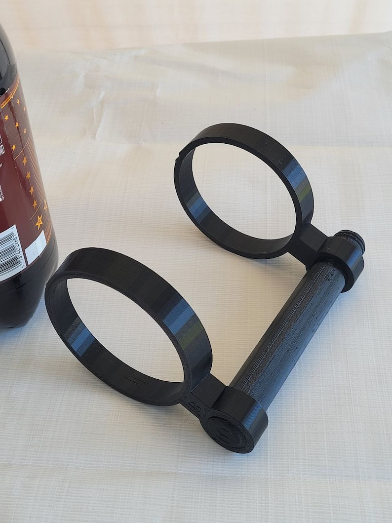 May include: A black plastic drink holder with two circular openings and a handle. The holder is designed to hold two cans or bottles.