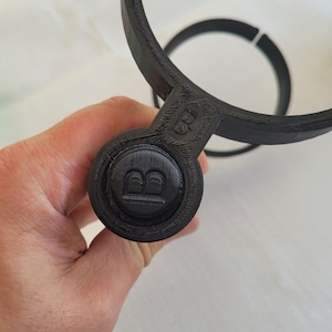 May include: Black plastic 3D printed cup holder with a round button on the side that has the letter "B" embossed on it.