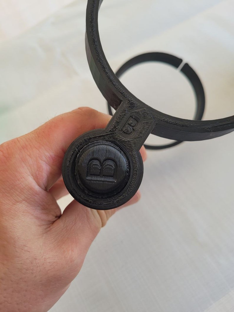 May include: Black plastic 3D printed cup holder with a round button on the side that has the letter "B" embossed on it.