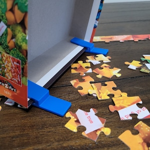 May include: Two blue plastic puzzle board clips hold the edges of a jigsaw puzzle box lid in place. The box lid has a colorful image of fortune cookies on it. The text "Cra-Z-Art" and "1671" are visible on the box. Scattered puzzle pieces are on a wooden table.