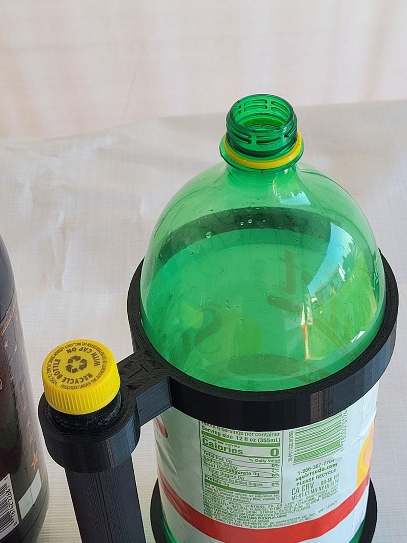 May include: A 3D printed bottle holder with a black base and a green plastic bottle inside. The bottle holder has a handle and a yellow cap on the top. The bottle has a label with nutritional information and a serving size of 12 fl oz (355mL).