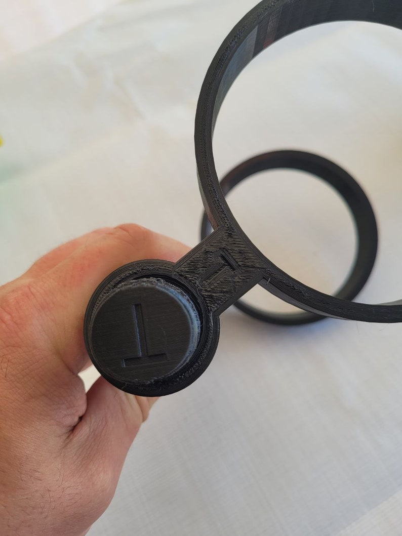 May include: Black plastic cup holder with a circular design. The holder has a small button on the side with the letter "L" printed on it.