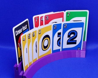 May include: A purple plastic card holder for playing Uno. The holder is curved and holds a variety of cards, including a "Draw Four" card, a "Wild" card, a "0" card, and several number cards.