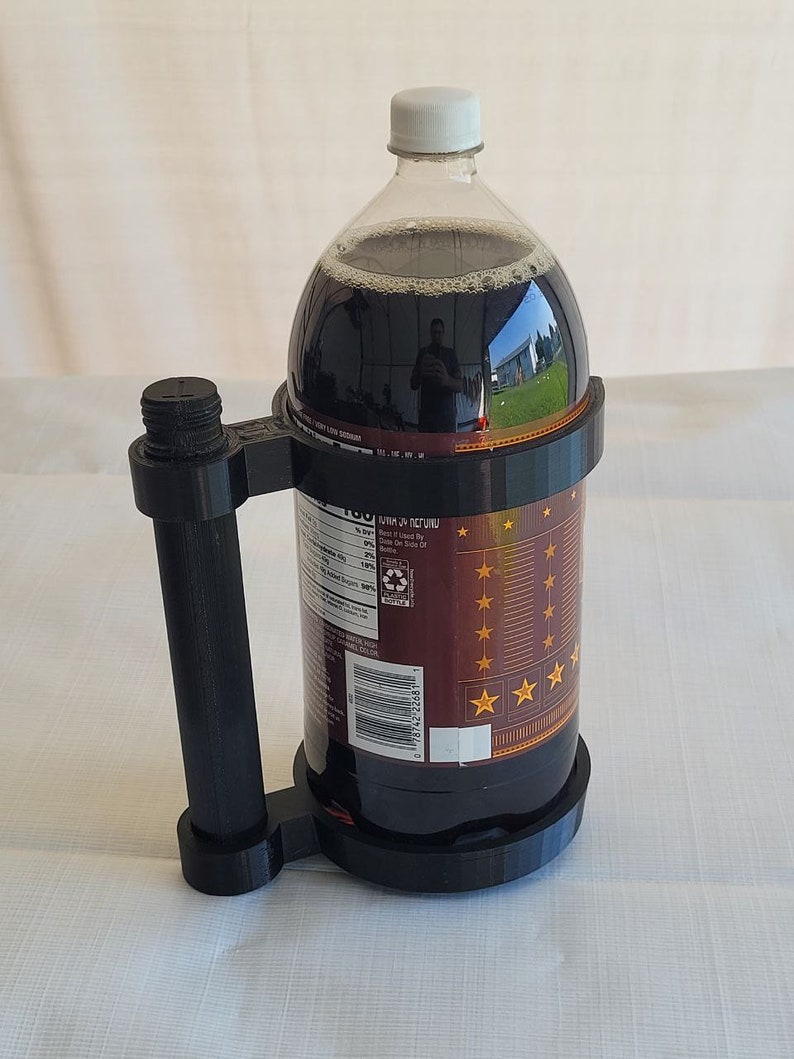 May include: A black 3D printed bottle holder with a handle. The holder is designed to hold a large plastic bottle of soda. The bottle is brown and has a white cap. The bottle label has a barcode and text that reads "100% of the Sugar is Naturally Occurring".