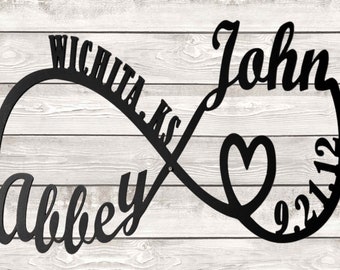 May include: Black metal infinity symbol with the names "Abbey" and "John" and the date "9.2.12" on a white wood background. A heart is in the center of the infinity symbol.