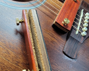 Könnte beinhalten: Eine Nahaufnahme eines hölzernen Gitarrenkapos mit zwei Messingknöpfen. Der Capo besteht aus mehreren Holzschichten und hat eine rotbraune Oberfläche.