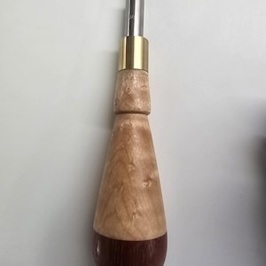 May include: A handcrafted screwdriver with a wooden handle. The handle is light brown with a dark brown base. A brass collar joins the handle to the silver metal shaft and the screwdriver bit.