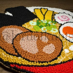 May include: A handmade, round rug depicting a bowl of ramen. The rug features various elements of ramen, including noodles, egg, seaweed, and meat. The colors include brown, yellow, white, black, red, and green.