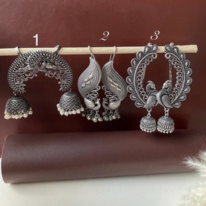 Pode incluir: Três pares de brincos de prata com designs intrincados. O primeiro par tem a forma de uma lua crescente com um charme de sino pendurado. O segundo par apresenta um design de pavão com sinos pendurados. O terceiro par é um design de pavão com dois pavões se olhando e sinos pendurados.