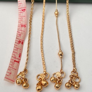 May include: Four gold-colored anklets with small bell charms are displayed. A pink and white measuring tape shows the length in inches. The anklets have different chain styles, including a delicate chain, a box chain, and a beaded chain.