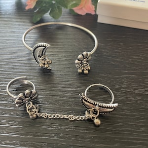 May include: A set of three silver-toned jewellery pieces: a bangle bracelet with a crescent moon and flower charm, and two finger rings connected by a chain. The rings feature a crescent moon and a flower charm.
