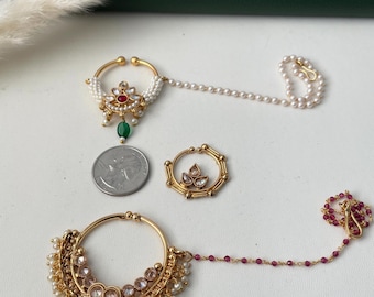 May include: Two gold-toned nose rings with chains and one smaller gold-toned nose ring. The top ring features white seed pearls, a central red stone, and a green drop. The bottom ring has clear stones and white pearls. A US quarter is shown for scale.