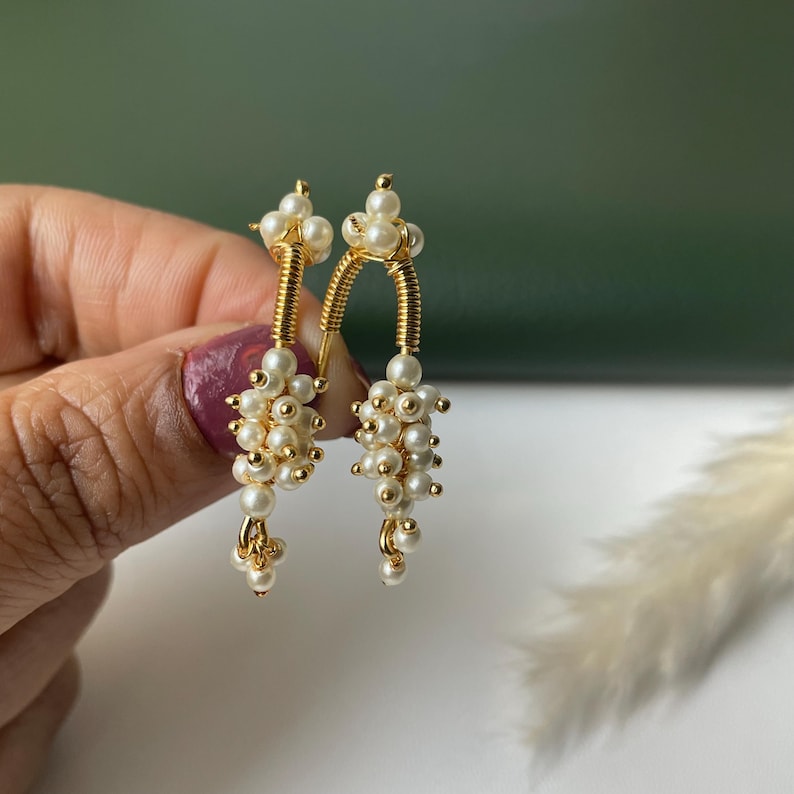 Pode incluir: Um par de brincos dourados com aglomerados de pequenas p&eacute;rolas brancas. Os brincos t&ecirc;m um design &uacute;nico com um elemento de fio enrolado e um aglomerado de p&eacute;rolas penduradas. O fundo &eacute; verde suave e branco.