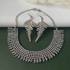 May include: Silver-coloured necklace and earring set. The necklace has a layered design with small, round beads and leaf-shaped pendants. The earrings are shaped like detailed fern leaves, creating a matching set.