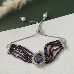 May include: A silver bracelet with a central, teardrop-shaped purple gemstone surrounded by small white stones. The bracelet has multiple strands of small, dark purple beads and adjustable pearl accents.