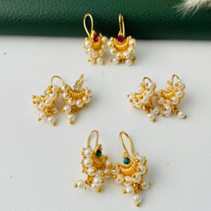 May include: Several pairs of gold-colored earrings with pearl and gemstone accents. The earrings feature various designs, including small red and green gemstones. The earrings are displayed on a white surface.