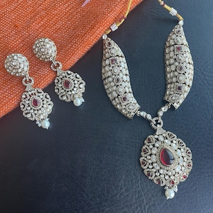 Puede incluir: Un conjunto de joyas plateadas con un collar y pendientes a juego. El collar tiene un colgante con una gran piedra roja en forma de lágrima, rodeada de piedras blancas más pequeñas. Los pendientes y el collar están adornados con detalles intrincados y piedras rojas más pequeñas.