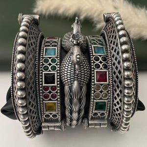 May include: Silver-toned peacock-themed bangles with colorful gemstone accents. The bangles feature intricate detailing, including a peacock design and geometric patterns. The gemstones are square and round, in various colors.