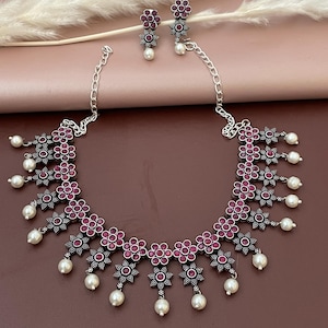 May include: A silver-toned necklace and earring set. The necklace features a floral design with red center stones and dangling faux pearls. The matching earrings have a similar floral design with a single pearl. The jewelry is displayed on a pink and brown background.