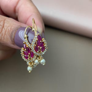 May include: Gold-coloured earrings with a teardrop design. Each earring features a cluster of oval-cut, ruby-coloured stones surrounded by small, clear crystals. Below the cluster hang three small, white pearls. The earrings are held by a hand.