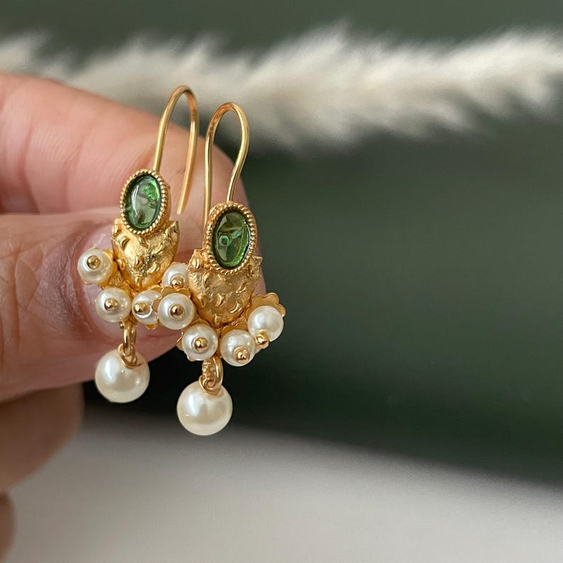 Pode incluir: Brincos dourados com pedras preciosas verdes ovais e aglomerados de pequenas p&eacute;rolas brancas redondas. Os brincos t&ecirc;m um design texturizado e ornamentado com fecho de gancho. Uma joia.